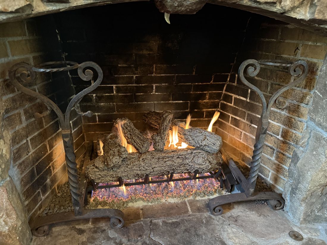 Gas logs burning in an old brick firebox with tall wrought iron andirons and decorative glass under the grate