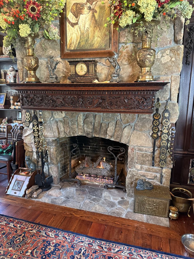 A stone fireplace with an ornately carved dark wood mantel, brass fittings and antiques, with gas logs burning