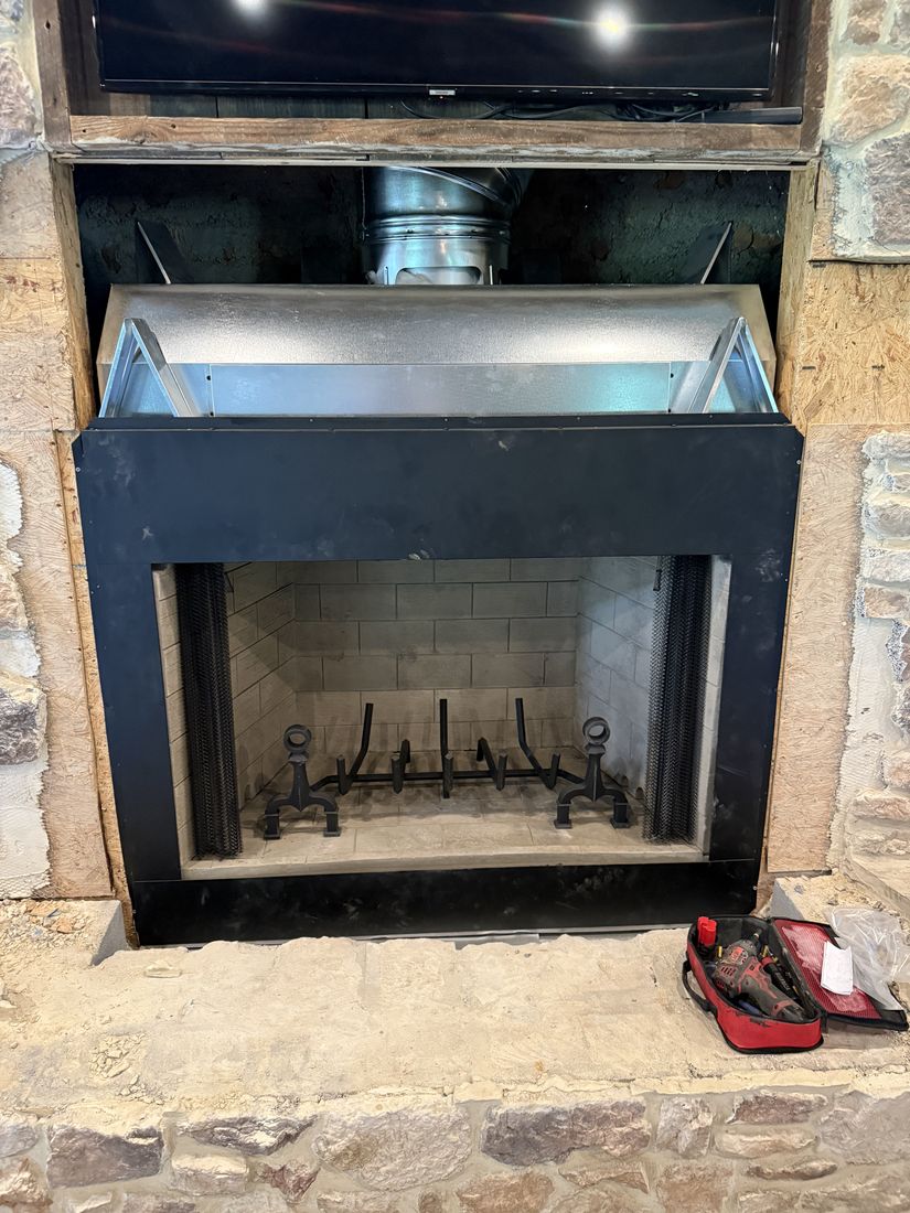 A fireplace unit part way into a stone opening with ductwork above it and a tool bag on the hearth