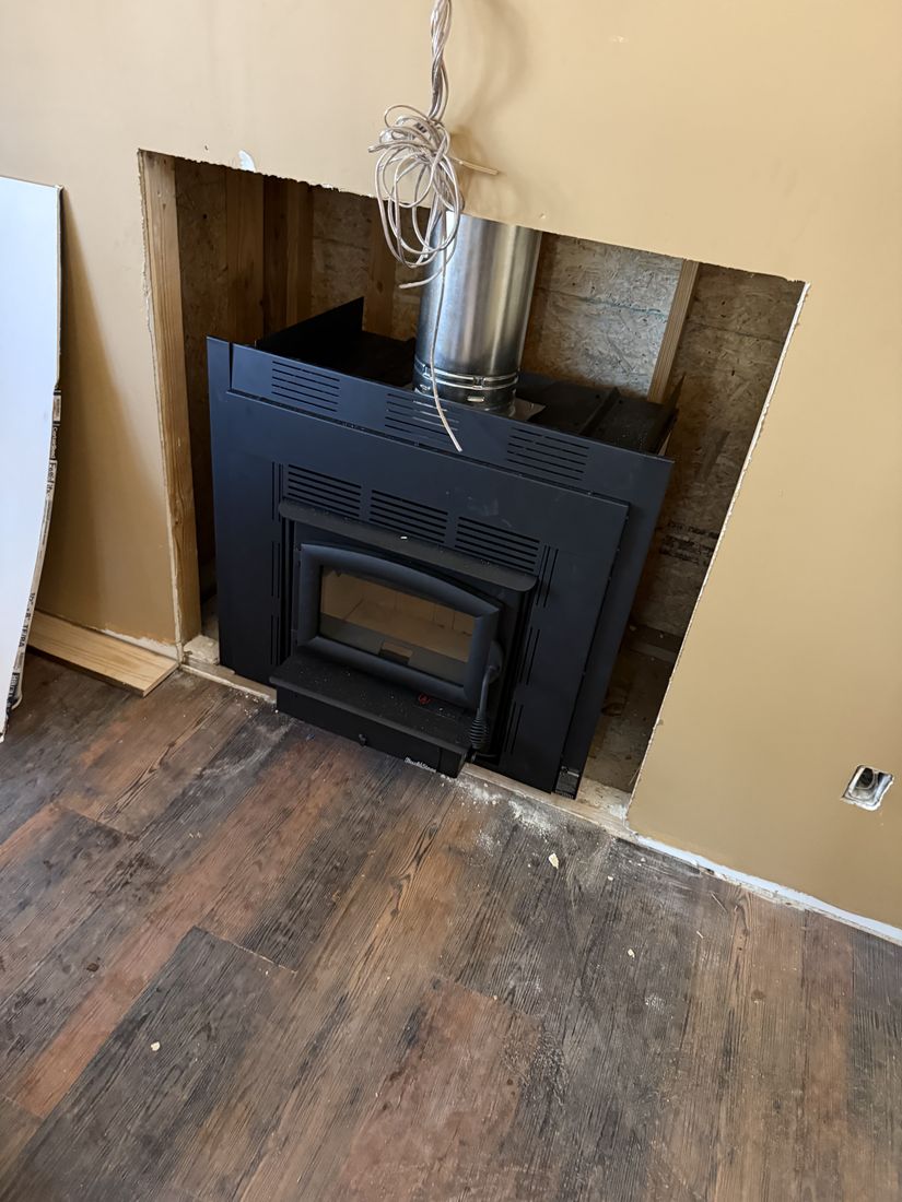 A black wood burning insert being fitted into an existing wall opening with a stainless steel liner running out of the top