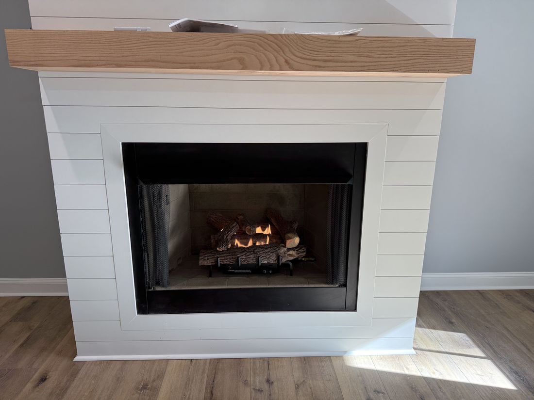 A white shiplap fireplace surround with a thick wood mantel and a gas log set burning behind the screen