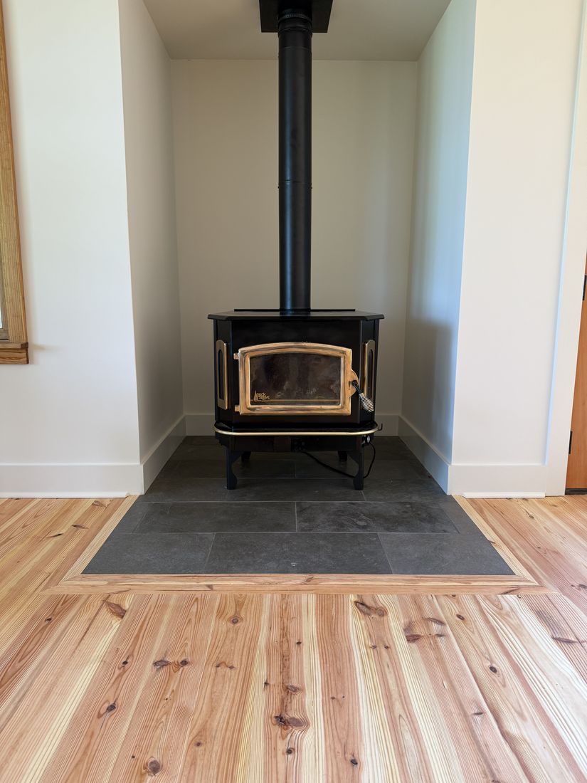 A freestanding wood stove with a brass trimmed door on a dark tile hearth pad set into wood flooring