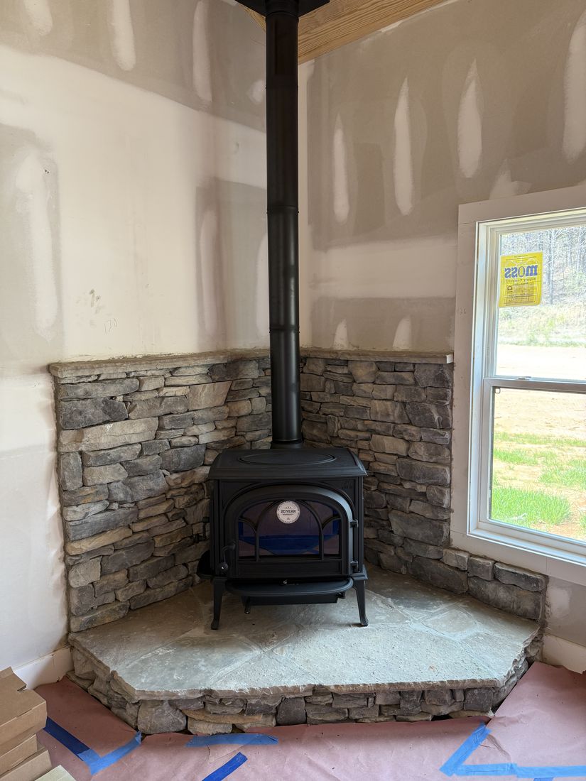 A black wood stove in a corner on a stone hearth pad with stacked stone behind it, room still under construction
