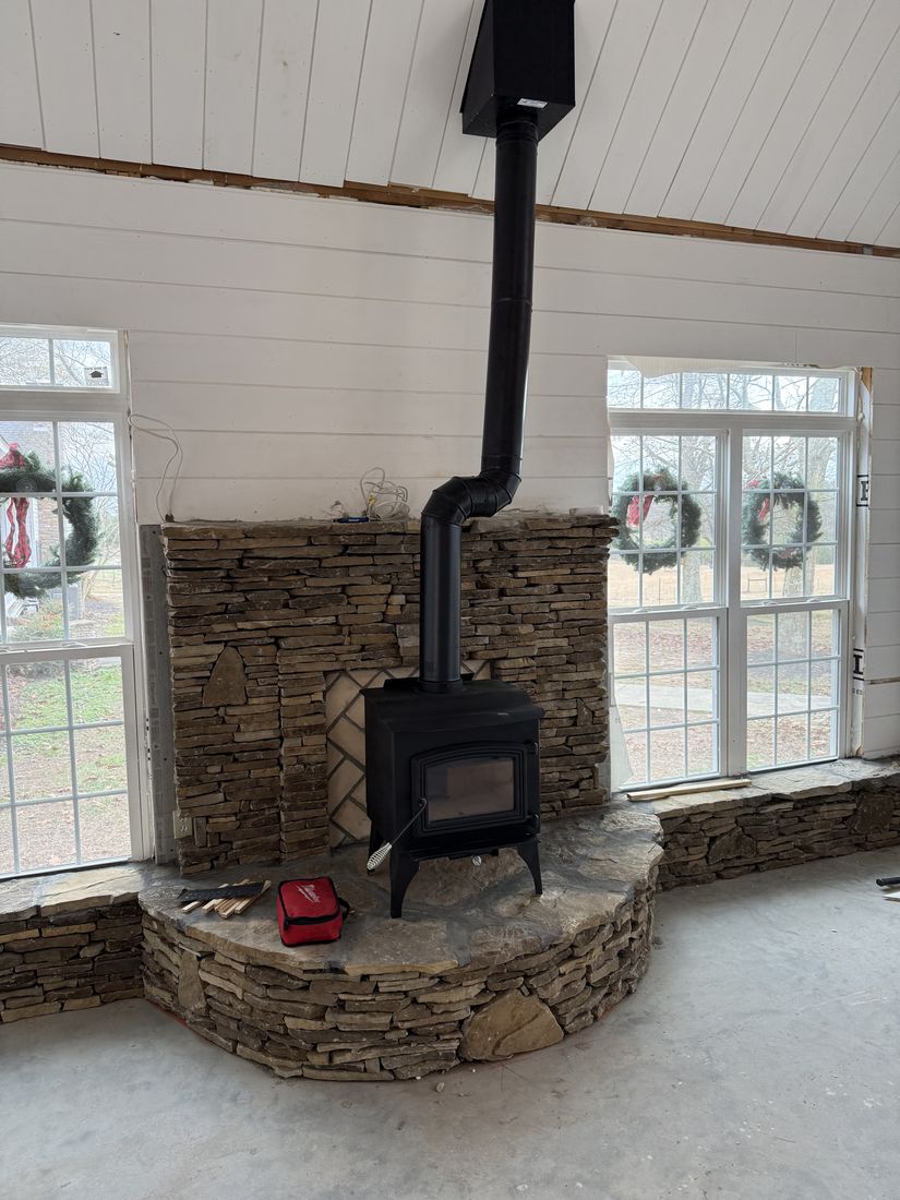 A black freestanding wood stove on a curved stacked stone hearth pad with a stone backing wall between two windows